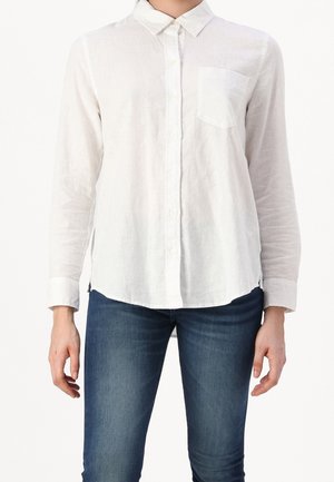 Personne portant une chemise blanche à manches longues boutonnée avec une poche poitrine et un jean bleu, debout devant un fond clair uni.