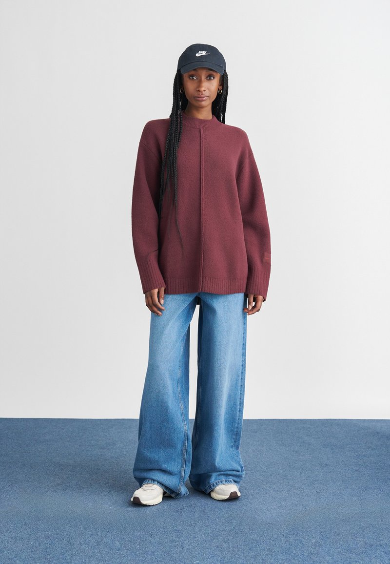 Bordeaux stickad tröja med högkrage och ribbat fåll; tillsammans med ljusblå vida jeans och vita sneakers. Svart Nike-keps på huvudet.