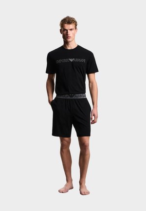 Giovane uomo in piedi a piedi nudi che indossa una T-shirt nera Emporio Armani e pantaloncini abbinati con logo sul petto e in vita su sfondo bianco.