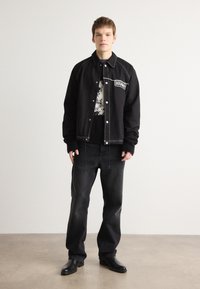 Giubbotto bomber nero con cuciture e logo bianchi, abbinato a jeans neri e stivali di pelle, su uno sfondo neutro.