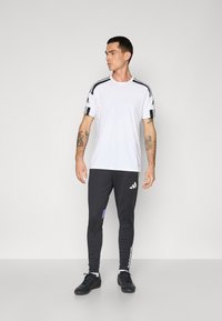 Witte sport T-shirt met zwarte schouderstrepen, gecombineerd met donkergrijze broek met kleurrijke accenten en zijstrepen, gedragen met zwarte schoenen.