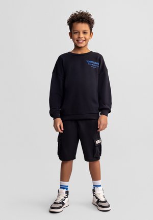 Ragazzo sorridente in piedi, con felpa nera con la scritta Tokyo Japan, pantaloncini cargo neri, sneakers bianche e calzini bianchi con strisce blu su sfondo grigio.