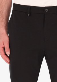 Pantaloni neri realizzati in un tessuto liscio, con frontale piatto, chiusura con bottone e passanti per la cintura, mostrati con una mano appoggiata di lato.