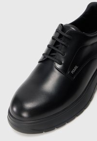 Chaussure habillée en cuir noir avec un bout rond, texture lisse, et six œillets pour les lacets ; présente un logo sur le côté.