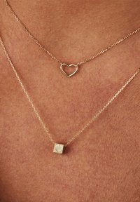 Layered gold necklaces featuring a heart pendant and a square charm with an engraved heart design, both on delicate chains.