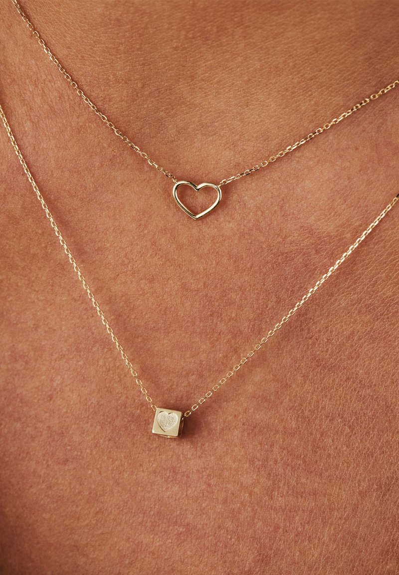 Layered gold necklaces featuring a heart pendant and a square charm with an engraved heart design, both on delicate chains.