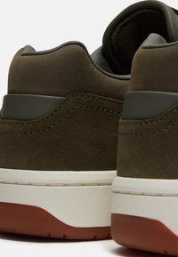 Primer plano de zapatillas de ante verde oscuro con entresuela blanca y suela exterior de goma marrón, mostrando el collar acolchado y costuras detalladas.