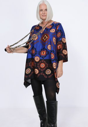 Tunique bleue à motifs bruns et orange, présentant une coupe ample, des manches courtes et un ourlet plus long à l'arrière. Portée avec des leggings noirs et des bottes noires longues et texturées.