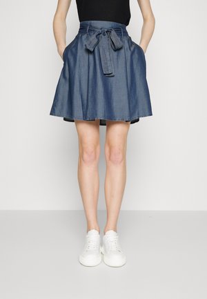 Femme portant une jupe en jean bleue longueur genou avec une ceinture nouée à la taille, un haut noir, des baskets blanches et les mains dans les poches de la jupe.