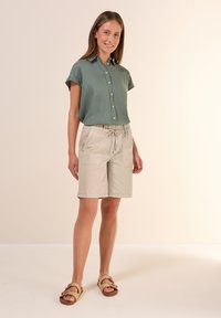 Kortärmad, knappad skjorta i grön tyg, tillsammans med beige shorts med elastisk midja och snörning. Modellen bär ljusbruna sandaler.