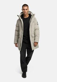 Jauns vīrietis staigā, ģērbts gaiši pelēkā kapucēta ziemas mētelī, melnā kreklā, melnās biksēs un melnās zābakos uz vienkrāsaina balta fona.