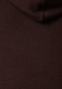 Bruine geribbelde stof met een zachte textuur en een capuchon. De verticale ribbels creëren een patroon dat de visuele diepte van het kledingstuk versterkt.