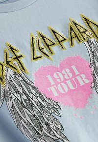 Jasnoniebieska koszulka z żółtym tekstem z cekinów "Def Leppard", różową grafiką serca z napisem "1981 TOUR" oraz czarnymi ilustracjami skrzydeł.