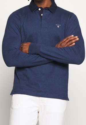 Homme portant un polo manches longues bleu marine avec le logo GANT, bras croisés, associé à un pantalon blanc sur un fond uni.