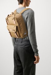 Brun ryggsekk med doble skulderstropper, topphåndtak, frontlomme og lærdetaljer. Laget av slitesterkt stoff med et minimalistisk design.