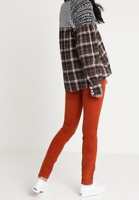 Femme portant un chemisier à manches longues à carreaux, un pantalon slim orange brûlé et des baskets blanches, debout avec un pied légèrement levé.