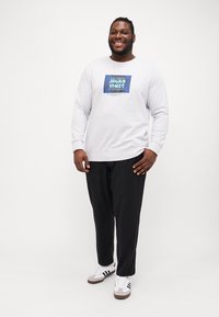 Helles graues Sweatshirt mit einem blauen Grafiklogo auf der Vorderseite, kombiniert mit schwarzen Hosen und weißen Sneakers mit schwarzen Akzenten.
