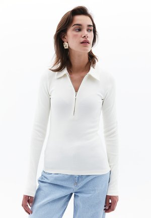 Witte lange mouwen top met een ritssluiting bij de kraag en ribbeltextuur, gecombineerd met lichtblauwe jeans. Een model toont de outfit tegen een witte achtergrond.