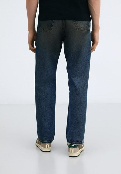 Jeans blu scuro con taglio dritto, con tasche posteriori visibili. Indossati con sneaker beige con dettagli verdi e marchio. Texture liscia.