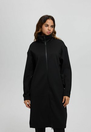 Kvinde med bølget hår iført en lang sort jakke med lynlås og høj krave, stående mod en ensartet lysegrå baggrund.