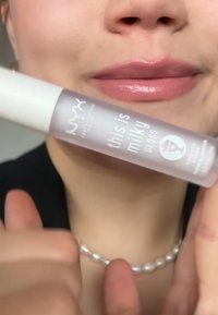 Gloss Milky NYX dans un tube transparent. L'étiquette présente un texte noir sur un fond givré. Les lèvres sont légèrement brillantes, et le cou est orné d'un collier de perles.