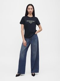 Femme debout, une main dans la poche, portant un t-shirt noir avec le texte "WEEKEND MaxMara 1983" et un jean bleu large sur fond blanc.