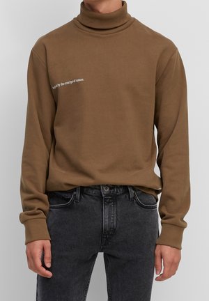 Sweat à col roulé marron avec le texte « Inspiré par le concept de la nature », porté avec un jean noir délavé, ne montrant que le torse et les bras.