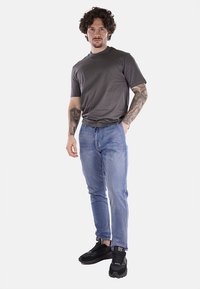 T-shirt grigio a maniche corte, jeans azzurri chiari e sneaker nere. I jeans sono attillati; il t-shirt ha un colletto rotondo. Tatuaggi visibili sulle braccia.