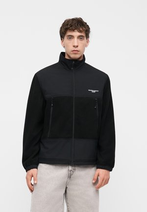 Man draagt een zwarte jas met ritssluiting, hoge kraag en twee voorzakken met rits, gecombineerd met lichtgrijze jeans, staand tegen een eenvoudige achtergrond.