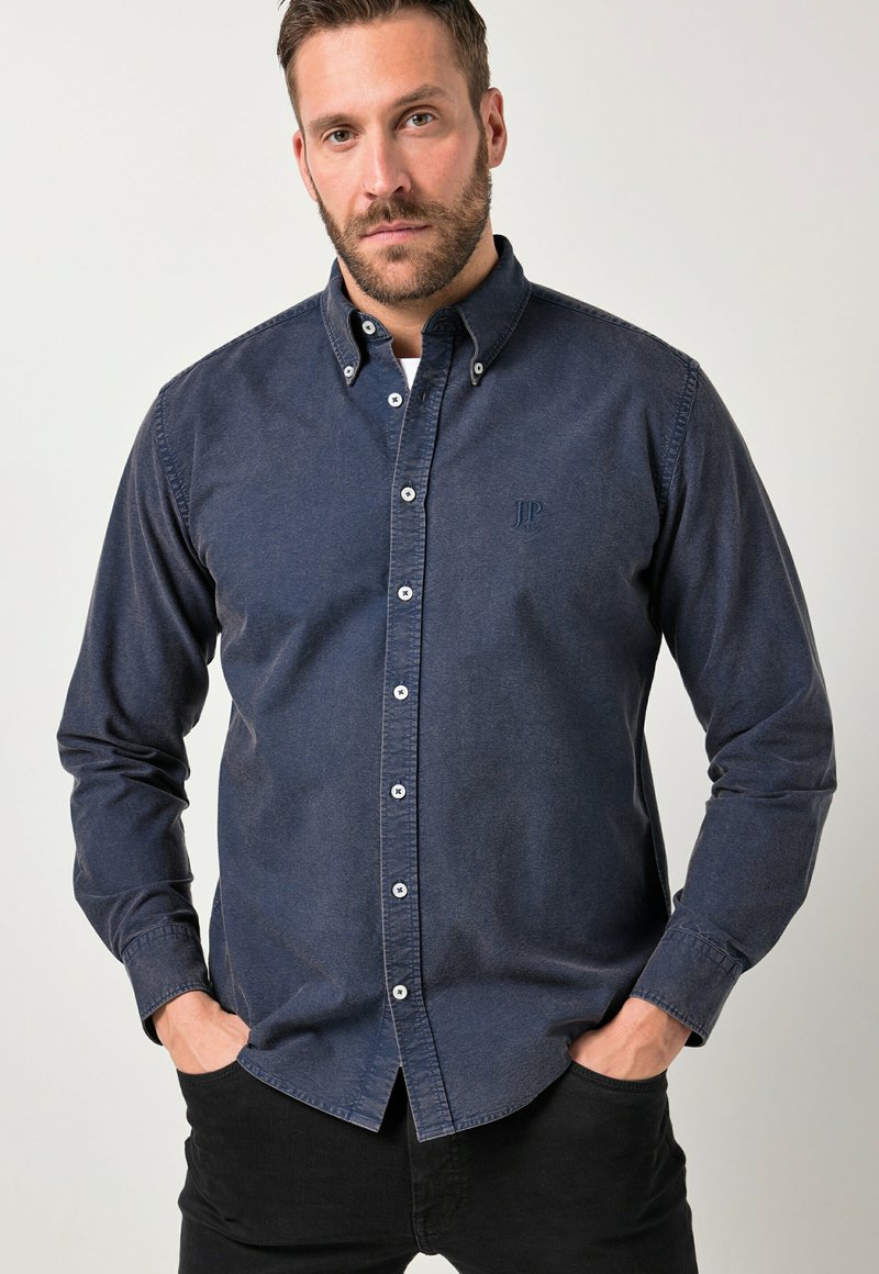 Uomo con la barba che indossa una camicia casual blu scuro abbottonata e pantaloni neri, in piedi con le mani in tasca contro uno sfondo semplice.