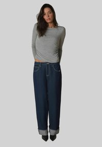 Langärmliges graues Oberteil kombiniert mit hoch taillierten, dunkelblauen Denim-Jeans mit aufgerollten Säumen und kontrastierenden Nähten. Schwarze, spitze Absätze inklusive.