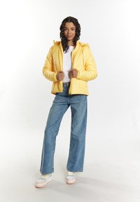 Giacca imbottita gialla con cappuccio, caratterizzata da chiusura frontale con zip e polsini a costine, abbinata a jeans larghi azzurri chiari e sneakers multicolori.