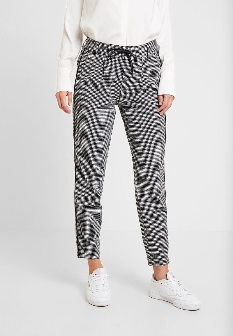 Pantalons à carreaux noir et blanc avec une taille à cordon, coupe ajustée, poches latérales et texture lisse, associés à des baskets blanches.