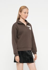 Žena s dlhými blond vlasmi, oblečená v hnedom svetri s polovičným zipsom a čiernej lesklej mini sukni s predným rozparkom, stojaca na bielom pozadí.