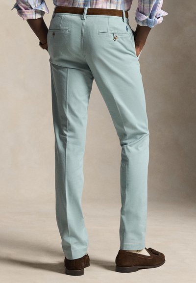 Vyras vilki šviesiai mėlynas siauras kelnes, rudos odos diržą, rudu zomšiniu batais be raištelių ir languotą marškinius su užriestomis rankovėmis, rankos kišenėse.