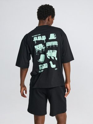 Osoba nosząca czarną koszulkę i szorty, na plecach koszulki znajdują się różne ilustracje jasnoniebieskich krzeseł i napis "THE SUNDAY CLUB."