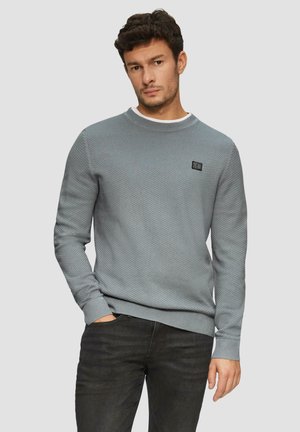 Helles graues strukturiertes Sweatshirt mit Rundhalsausschnitt, versehen mit einem schwarzen Logo auf der Brust sowie gerippten Bündchen und Saum. Kombiniert mit dunkler Jeans.