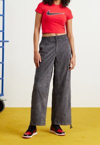 Czerwony krótki top z czarnym logo Nike, szare spodnie o szerokich nogawkach z teksturowanej tkaniny i kieszeniami, w zestawie z czarno-czerwonymi butami sportowymi.