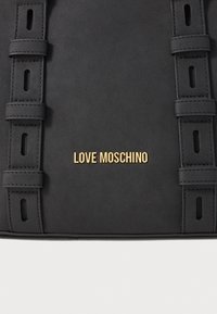 Crna torba Moschino sprijeda s zlatnim logotipom i dekorativnim šavovima na trakama s izrezbarenim detaljima.