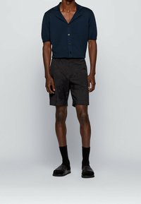 Tmavě modrá košile s krátkými rukávy a knoflíky, černé šortky, černé ponožky a nazouvací boty. Košile má límec a volný střih.