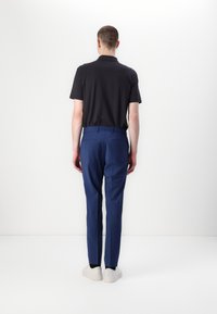 Schwarzes Poloshirt mit kurzen Ärmeln, blaue maßgeschneiderte Hosen und hellfarbige Sneakers, von hinten vor einem weißen Hintergrund betrachtet.