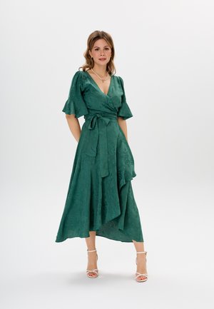 Abito verde avvolgente con maniche corte a volant, tessuto texturizzato, orlo alto-basso e vita legata, abbinato a sandali bianchi con cinturini.