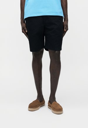 Persona con pantalones cortos negros, camisa azul claro y zapatos marrones sin cordones, de pie contra un fondo blanco liso.