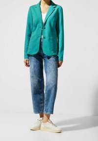 Blazer sarcelle à manches longues, col cranté, deux poches avant, porté avec un jean bleu clair et des baskets beiges. Tissu lisse, coupe ajustée.