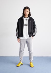 Črna jakna z zadrgo, s črtastimi belimi in zelenimi rokavi, čez belo majico, sive hlače in rumeni supergi z modrimi poudarki.