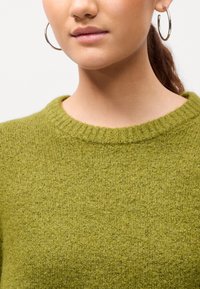 Zoomirane na osebo, ki nosi zeleno teksturirano pulover in velike srebrne obroče v ušesih, z lasmi zvezanimi nazaj.