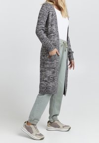 Lång, öppen-fram cardigan i en strukturerad grå stickning, med två sidofickor och en knappdetalj. Tillsammans med lätta gröna byxor och sneakers.