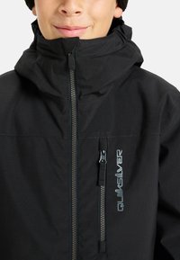 Zbliżenie na osobę w czarnej kurtce Quiksilver z wysokim kołnierzem i zapinaną na zamek kieszenią na piersi.