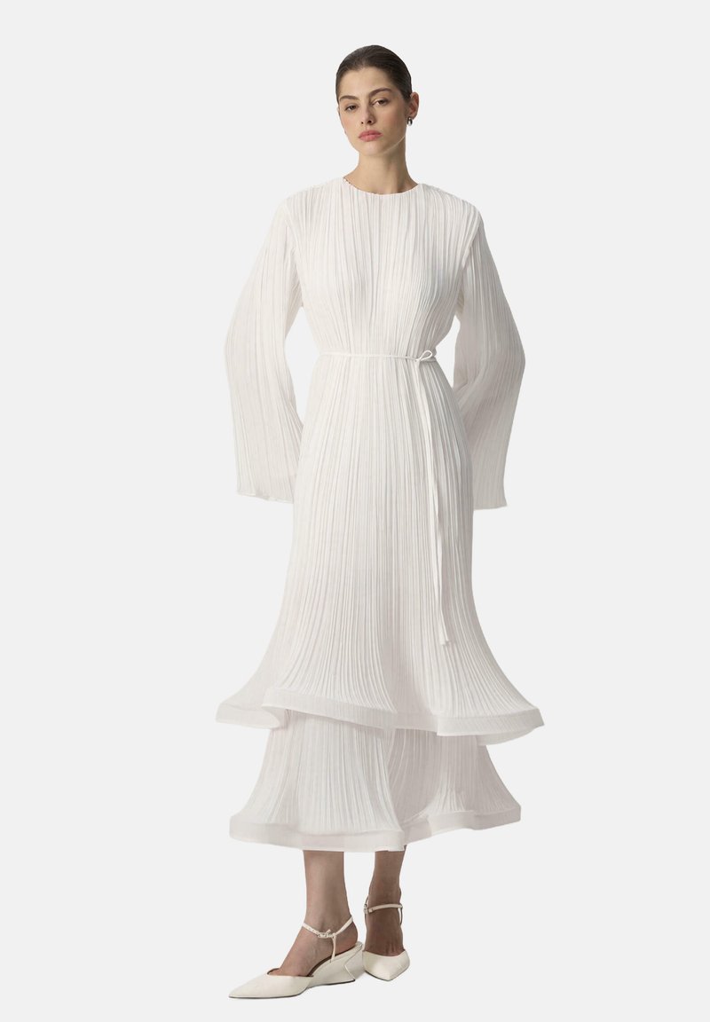 Vrouw in een wit geplooide midi-jurk met lange klokmouwen, een taillekoord en witte enkellaarsjes met puntige neus, staand tegen een effen achtergrond.