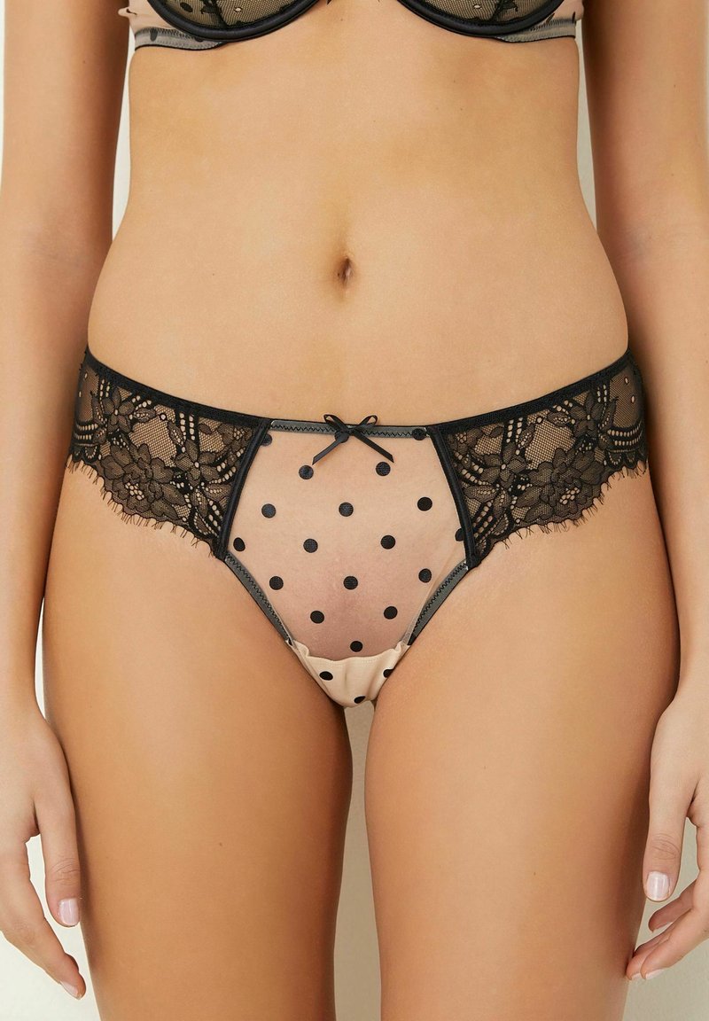 Spitzen- und Netzbikini mit schwarzen Punkten auf einem nude Hintergrund, versehen mit einem filigranen schwarzen Spitzenrand und einem kleinen Schleifchen vorne.